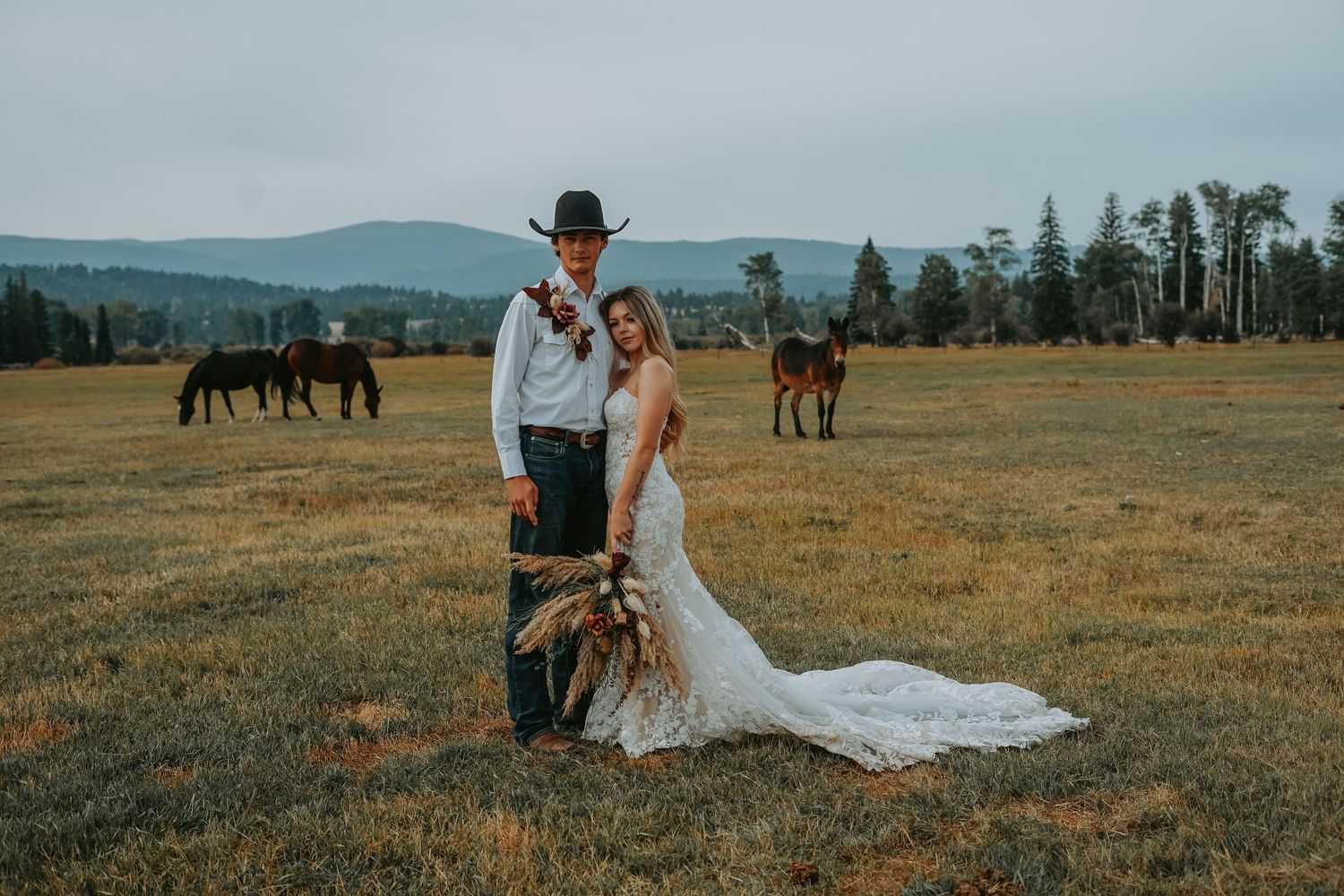 A western-themed wedding couple stands in a field with horses grazing in the mountainous background at sunset.