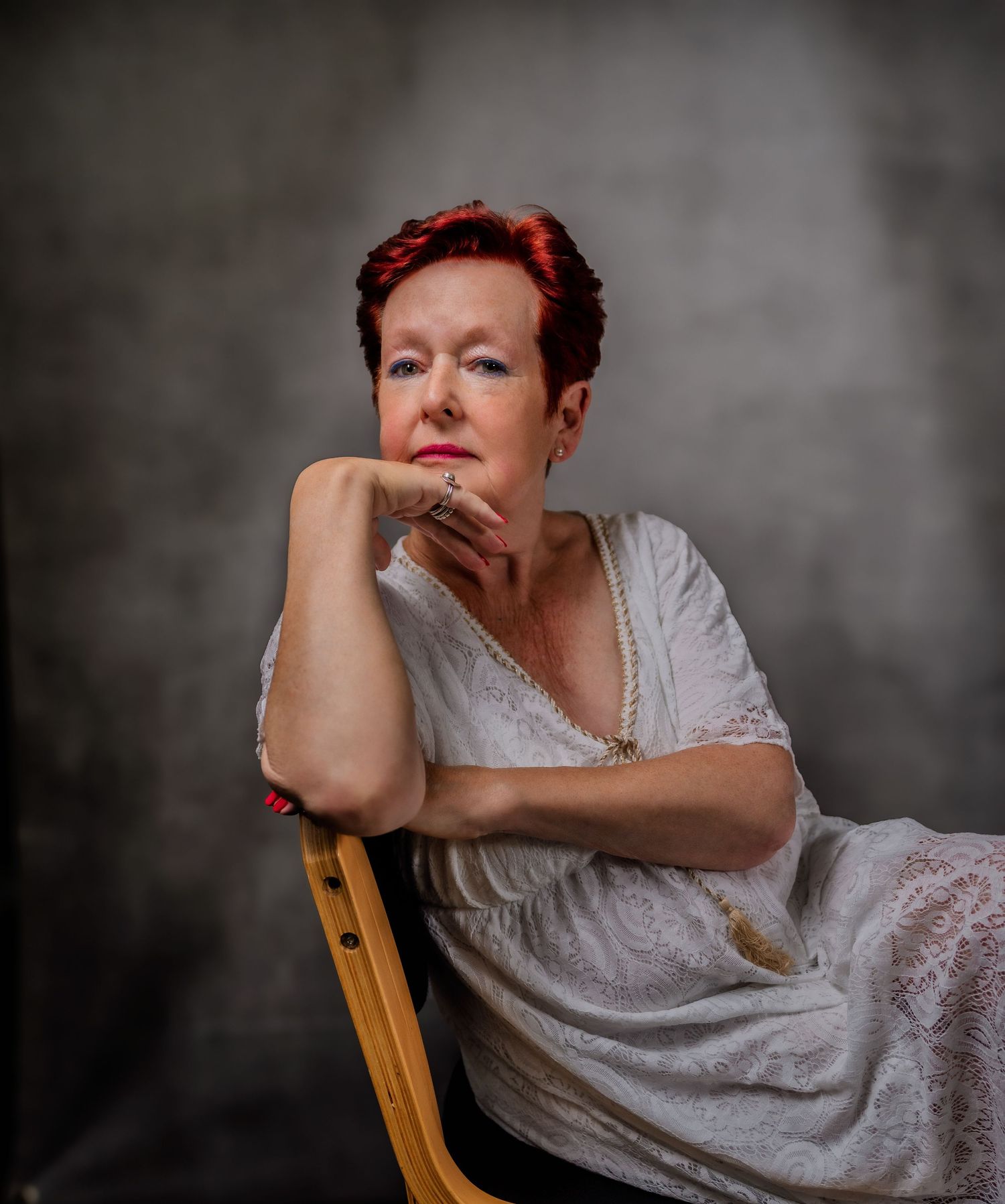 A contemplative figure in a white lace dress sits on a wooden chair against a dark backdrop.