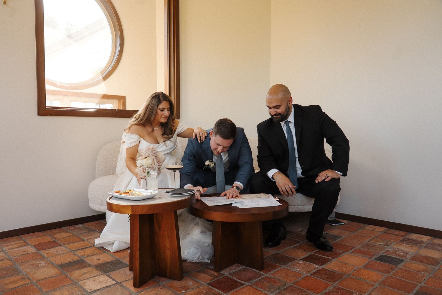 People gather around a small wooden table signing documents in a room with a circular window and terracotta tile floor.