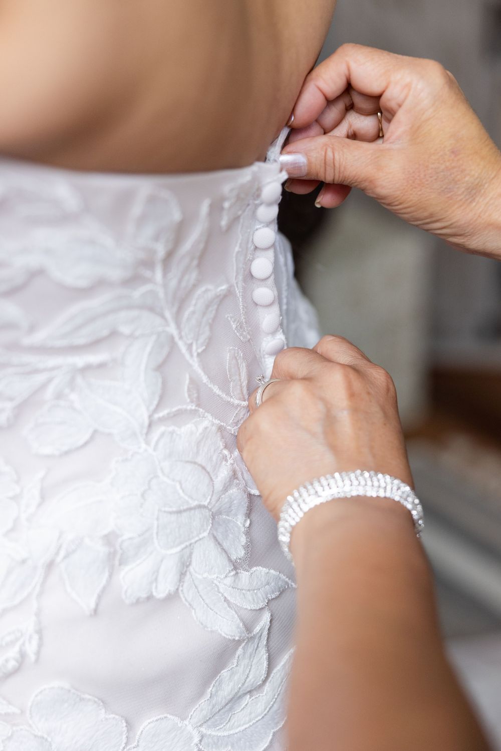Hands buttoning up the back of a delicate lace wedding dress with a pearl bracelet visible.