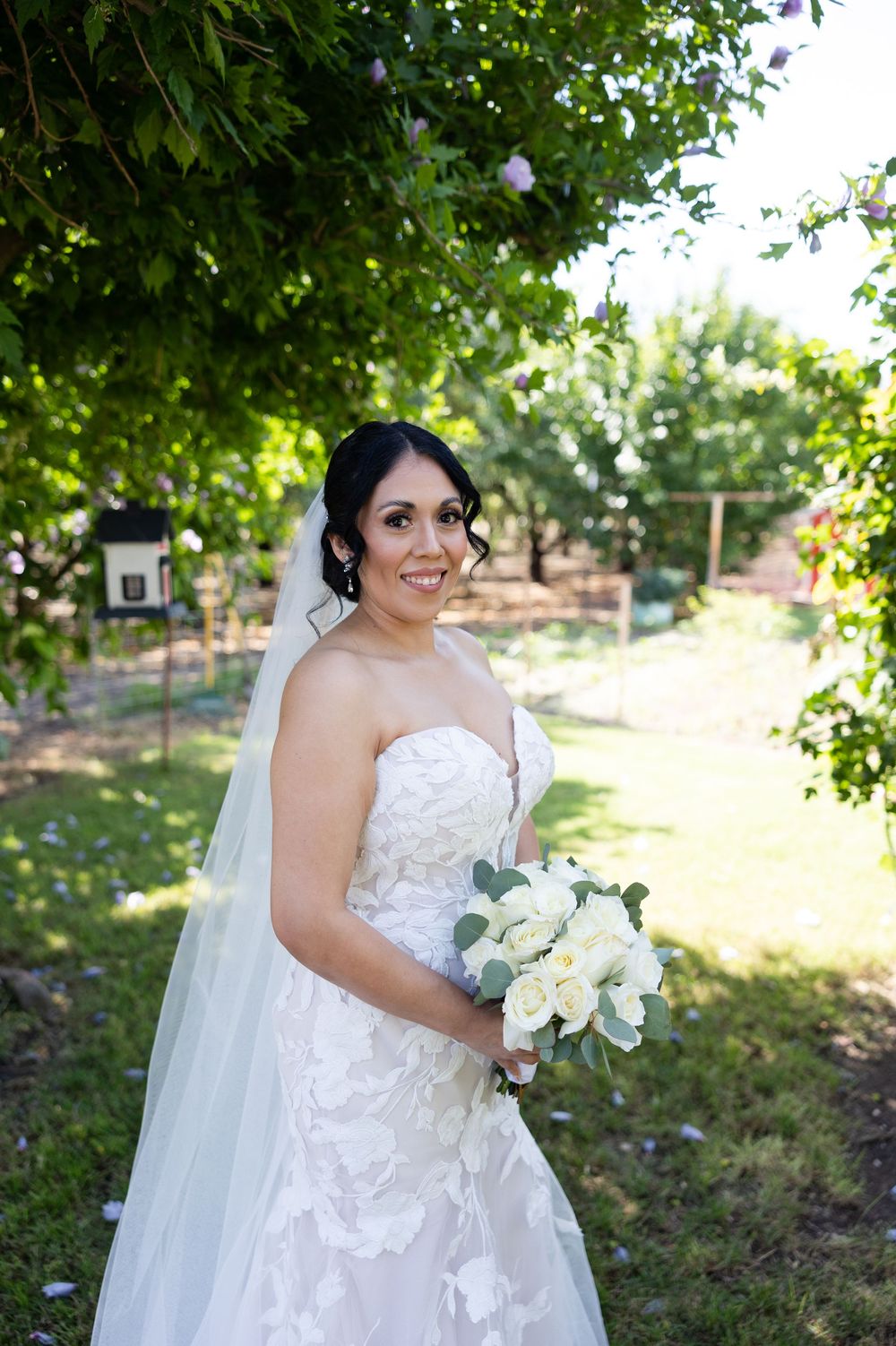 A bride holding white bouquet poses in a garden setting wearing a lace wedding gown.