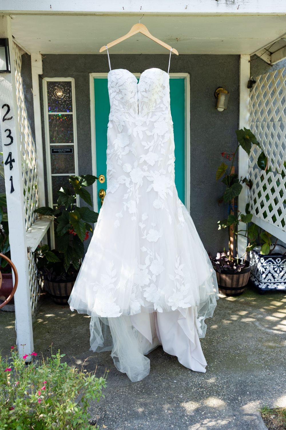 A beautiful white lace wedding dress hangs on display outside a home entrance with greenery.