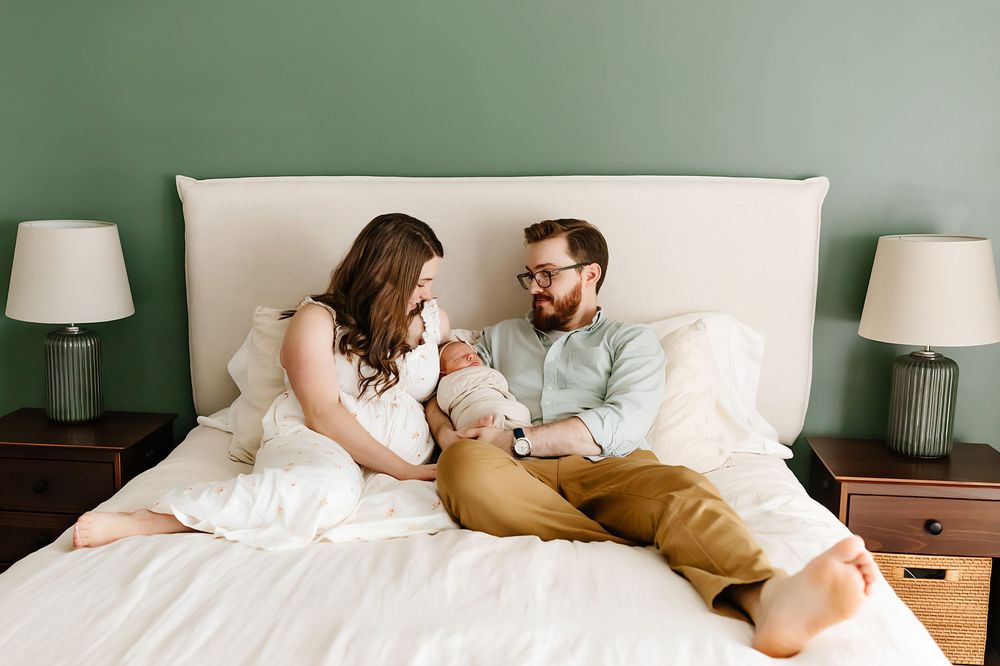 A couple sits on their bed with table lamps and green walls in a relaxed at-home portrait session.