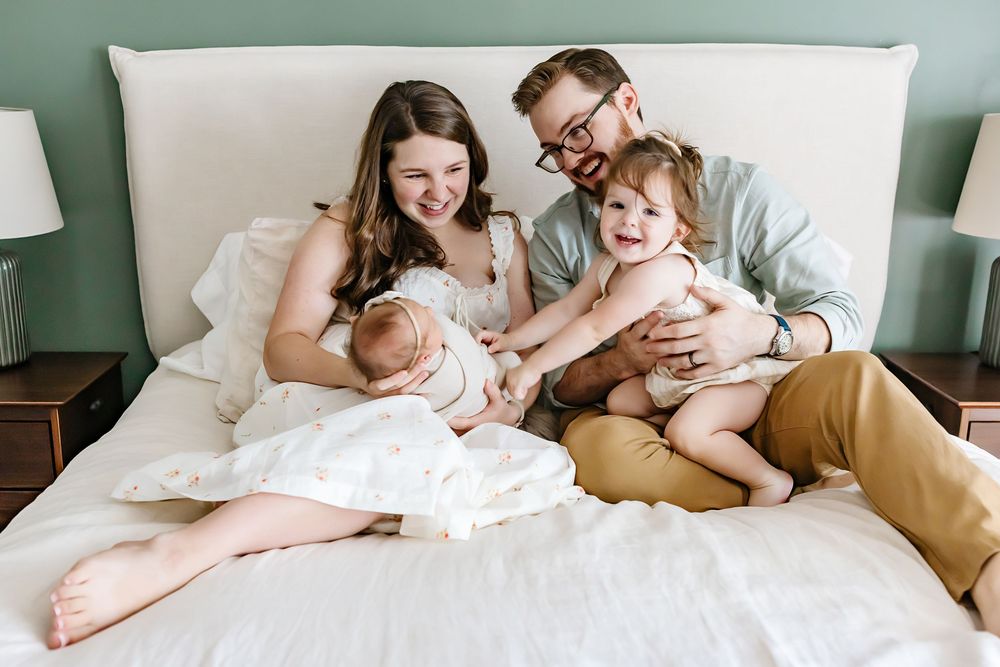 A family relaxes together on a white bed against a green wall in a candid lifestyle portrait.