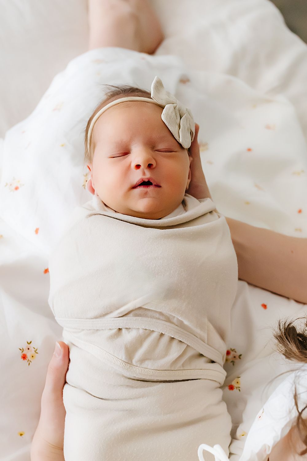 A sleeping newborn baby in cream colored wrap and bow headband rests peacefully on floral bedding.