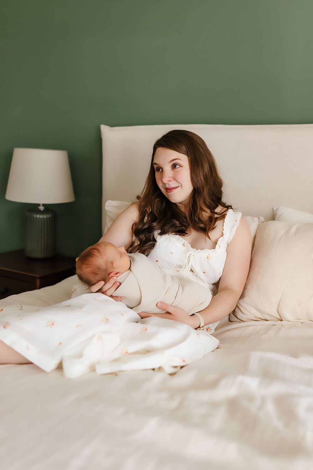 A mother and newborn baby rest together in a cozy bedroom with sage green walls and white bedding.