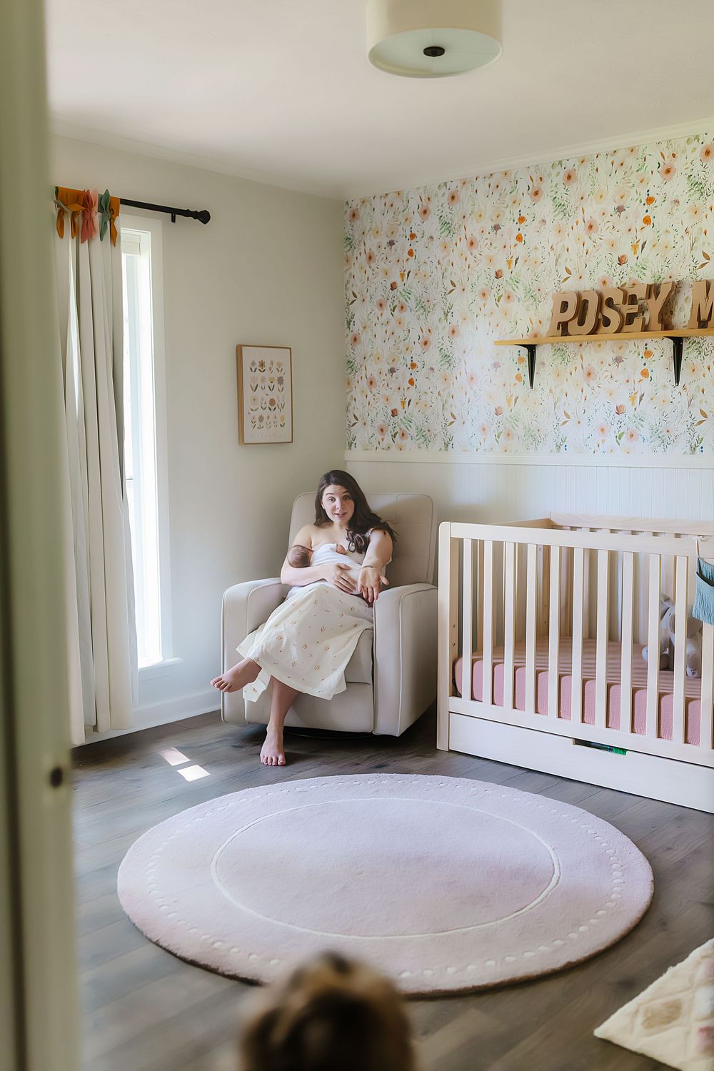 A nursery room features floral wallpaper, white crib, round pink rug and modern decor with natural lighting from window.