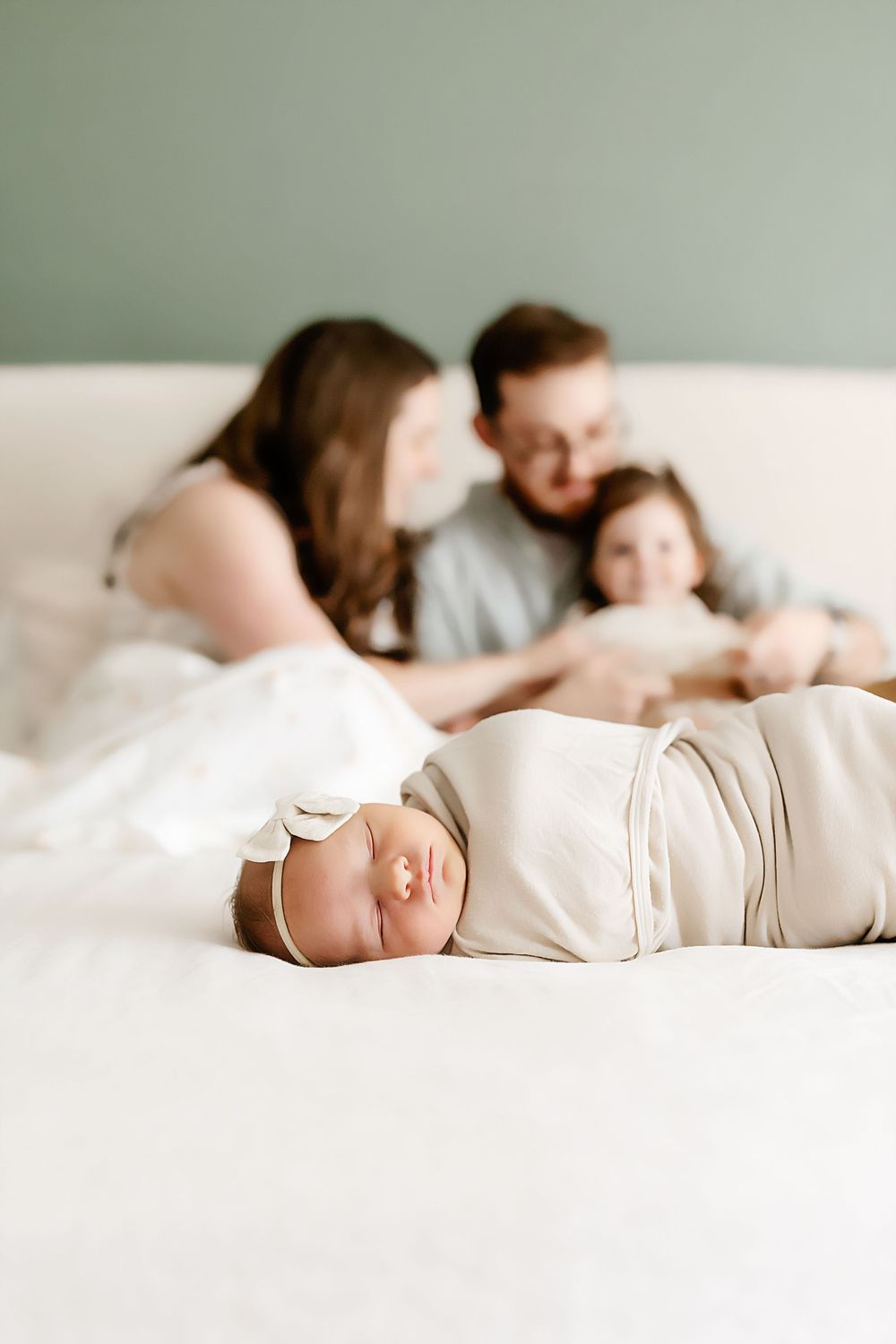 A sleeping newborn baby in soft white swaddle lies on a bed while a family gathers in the background.