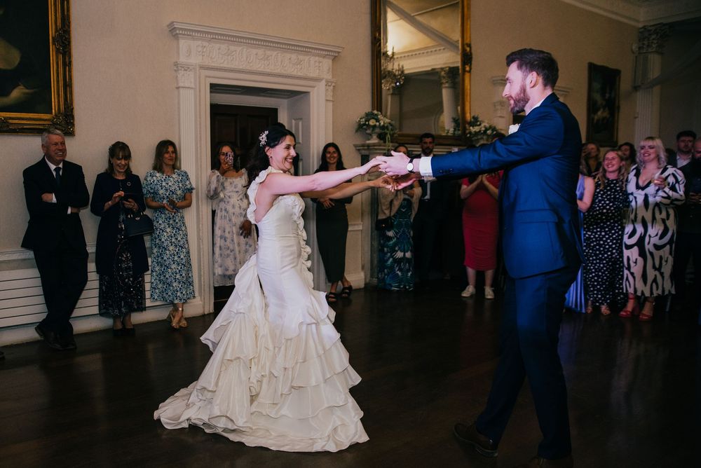 Bride in ruffled wedding dress dances with groom in blue suit while wedding guests watch at an elegant venue.