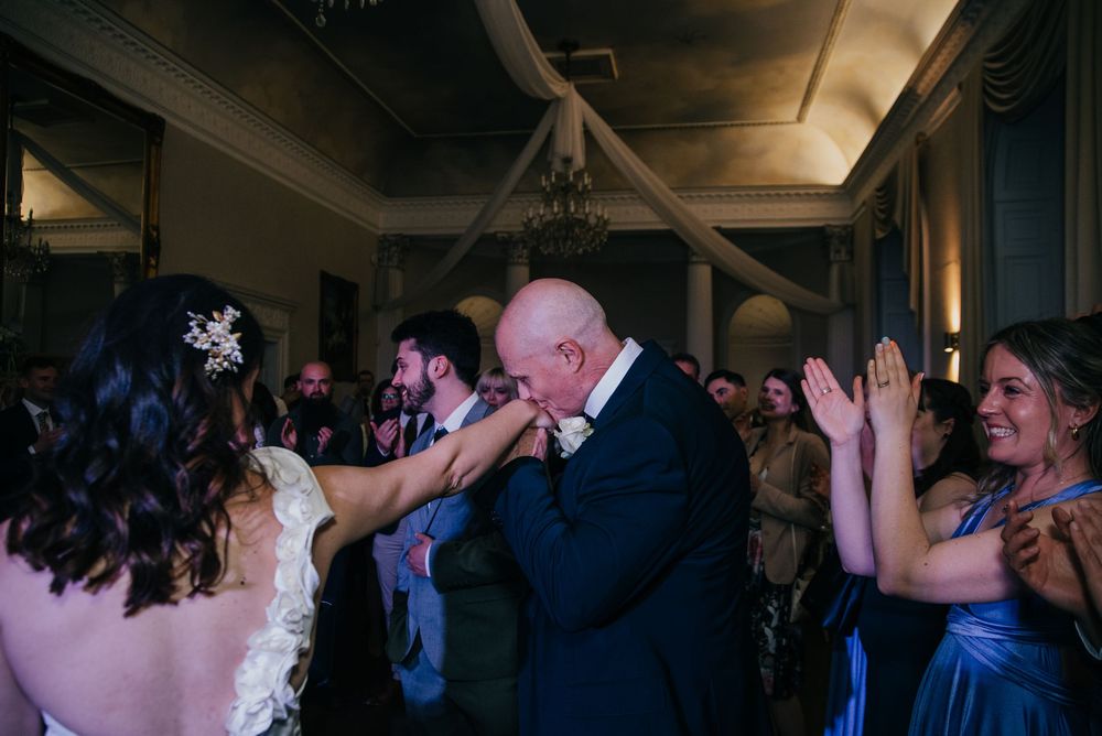 People dance and celebrate at a dimly lit indoor wedding reception with colorful lighting and festive atmosphere.