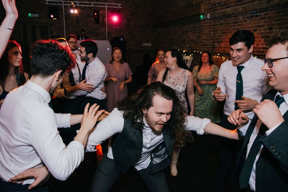 Wedding guests form a circle on the dance floor in a dimly lit venue with brick walls and soft lighting.
