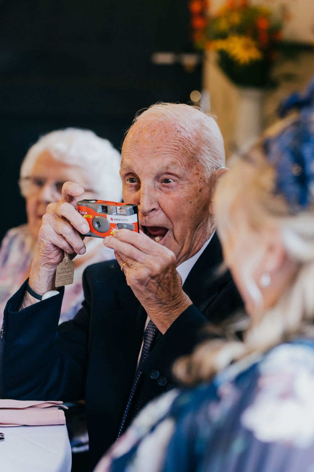 Senior dressed in dark suit takes photo with small camera at social gathering event.