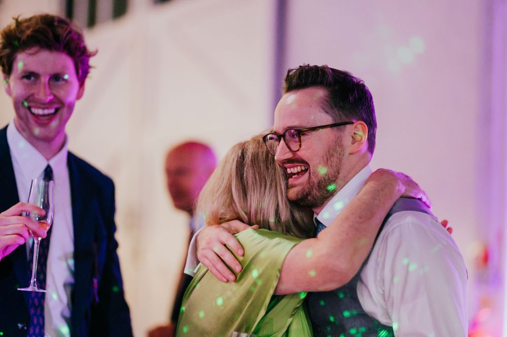 Wedding guests share laughter and embrace during a reception celebration with soft purple lighting.