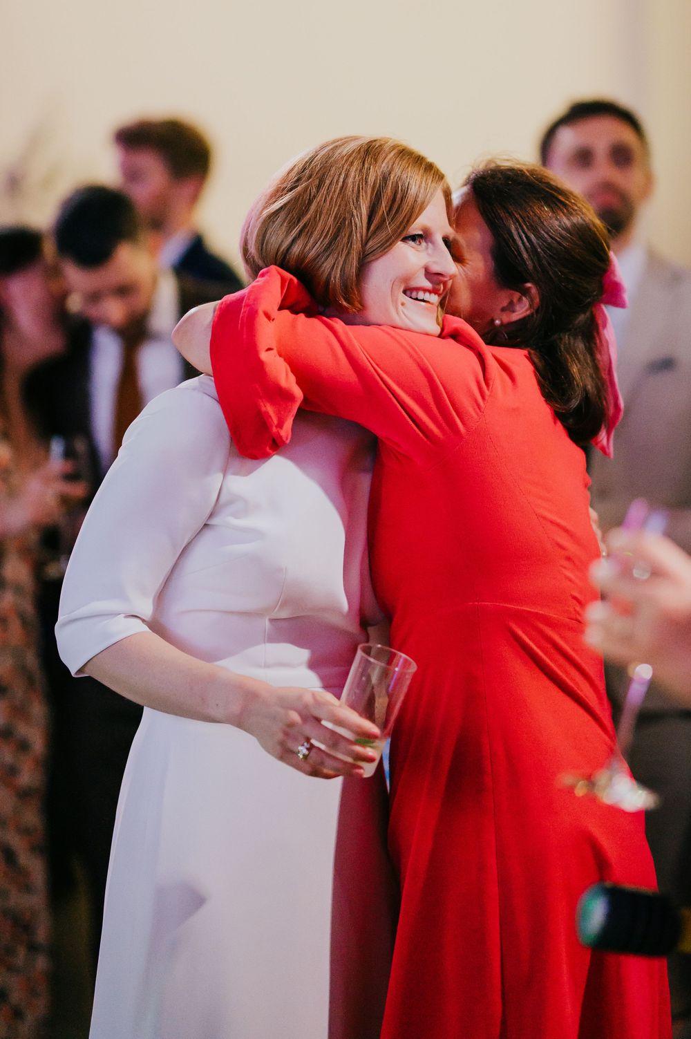 Two people share a warm embrace at a social gathering, with one wearing a white dress and the other in bright red attire.