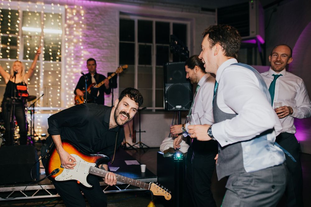 Live band performs energetically at wedding reception with guitarist and singer on dance floor with string lights backdrop.