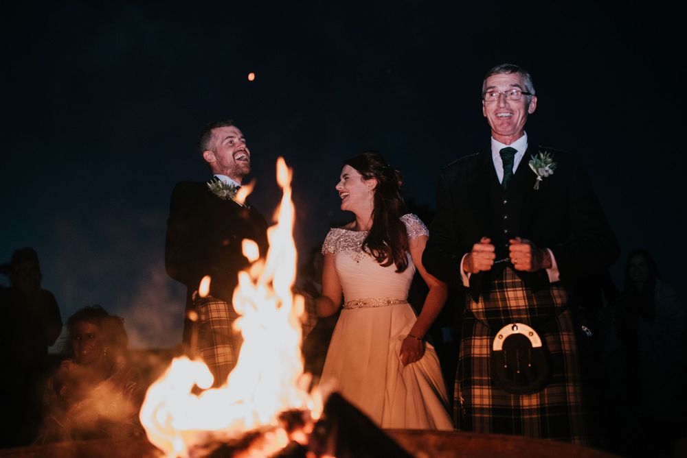 Three people gather around a bright bonfire at night, with one wearing traditional Scottish kilt attire.