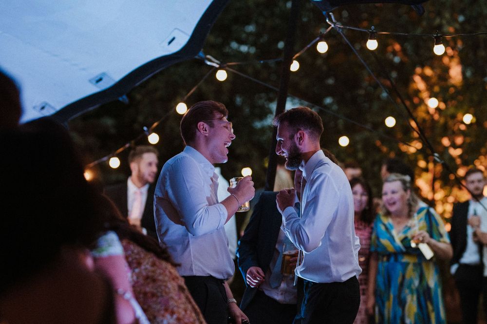 Wedding guests celebrating at an outdoor evening reception with string lights hanging overhead while enjoying drinks.