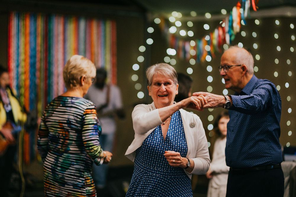 Elderly people dancing together at an indoor party decorated with sparkling string lights in the background.