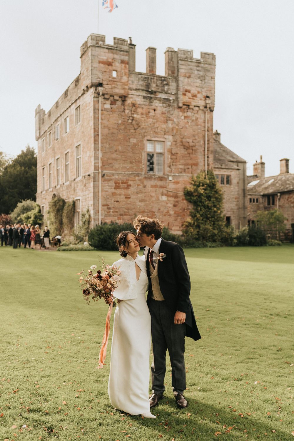 A couple in wedding attire stands on a manicured lawn in front of a historic stone castle on a sunny autumn day.