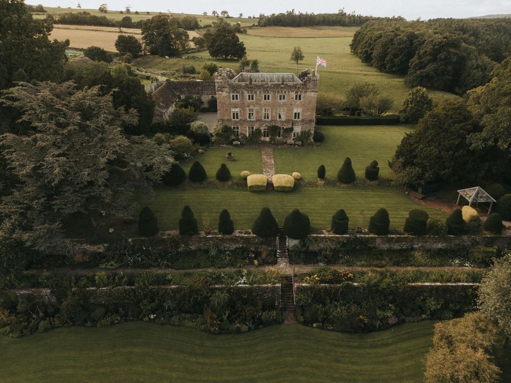A scenic aerial view of a rolling countryside estate with manicured gardens and stone buildings nestled among green hills.