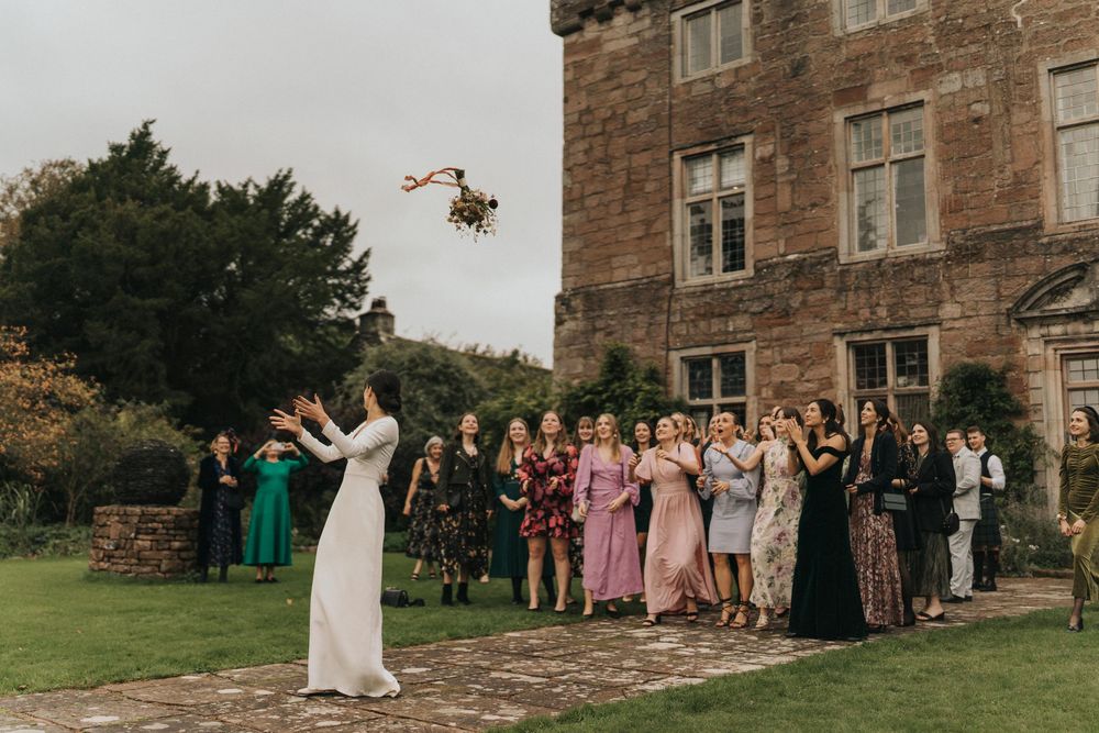 Wedding guests gather on a lawn outside a stone castle as someone tosses a bouquet into the air during the celebration.