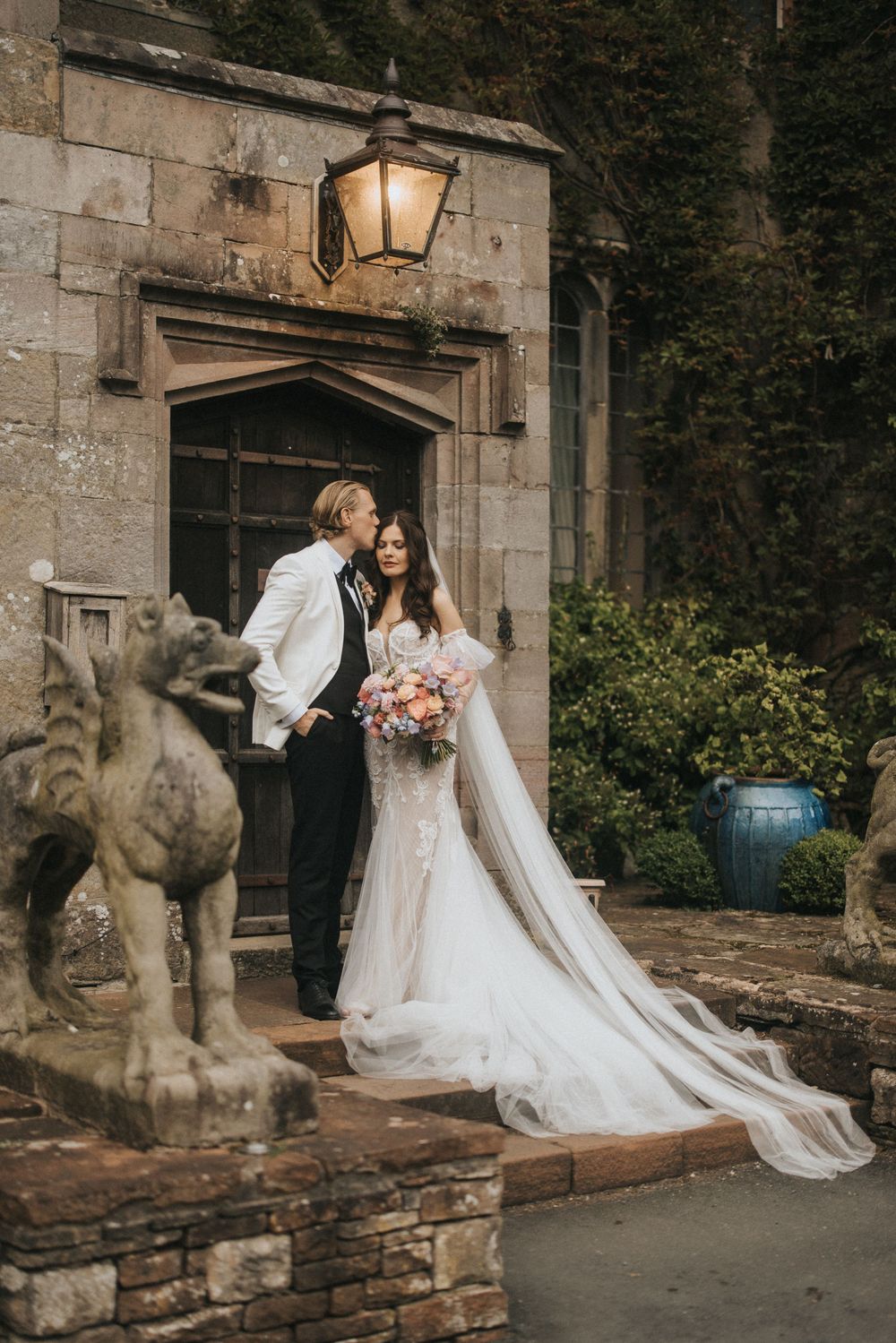 Romantic wedding couple poses in front of historic stone building with dramatic flowing white gown and bouquet.