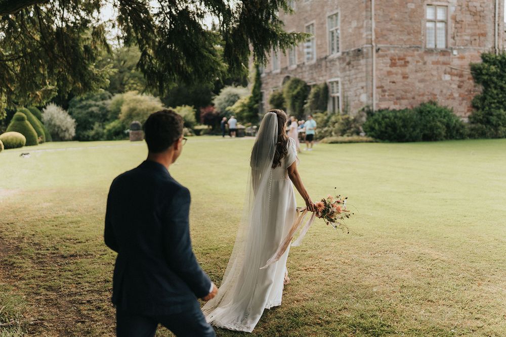A bride and groom walk together across a lush green lawn at their castle wedding venue.