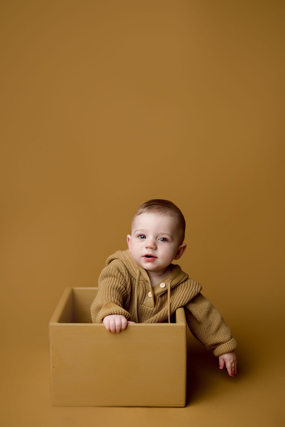 Baby in brown sweater sits inside cardboard box against warm brown backdrop in creative portrait series.