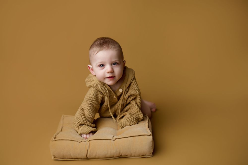 Baby in tan hoodie sits on beige cushion against warm brown studio backdrop.