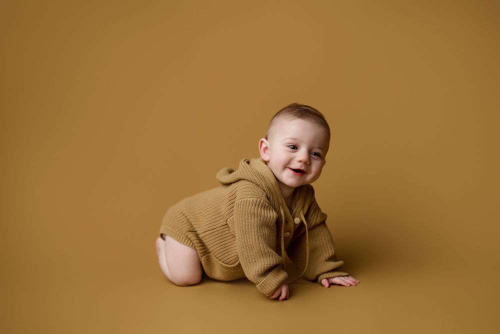 Baby wearing knitted tan sweater crawls on golden brown backdrop during studio photo session.