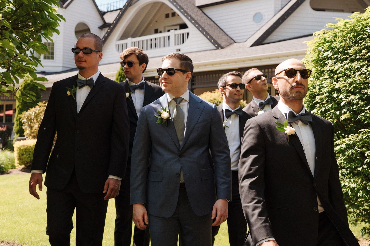 Group of groomsmen in formal attire with sunglasses pose together on a sunny day outside an elegant white mansion.