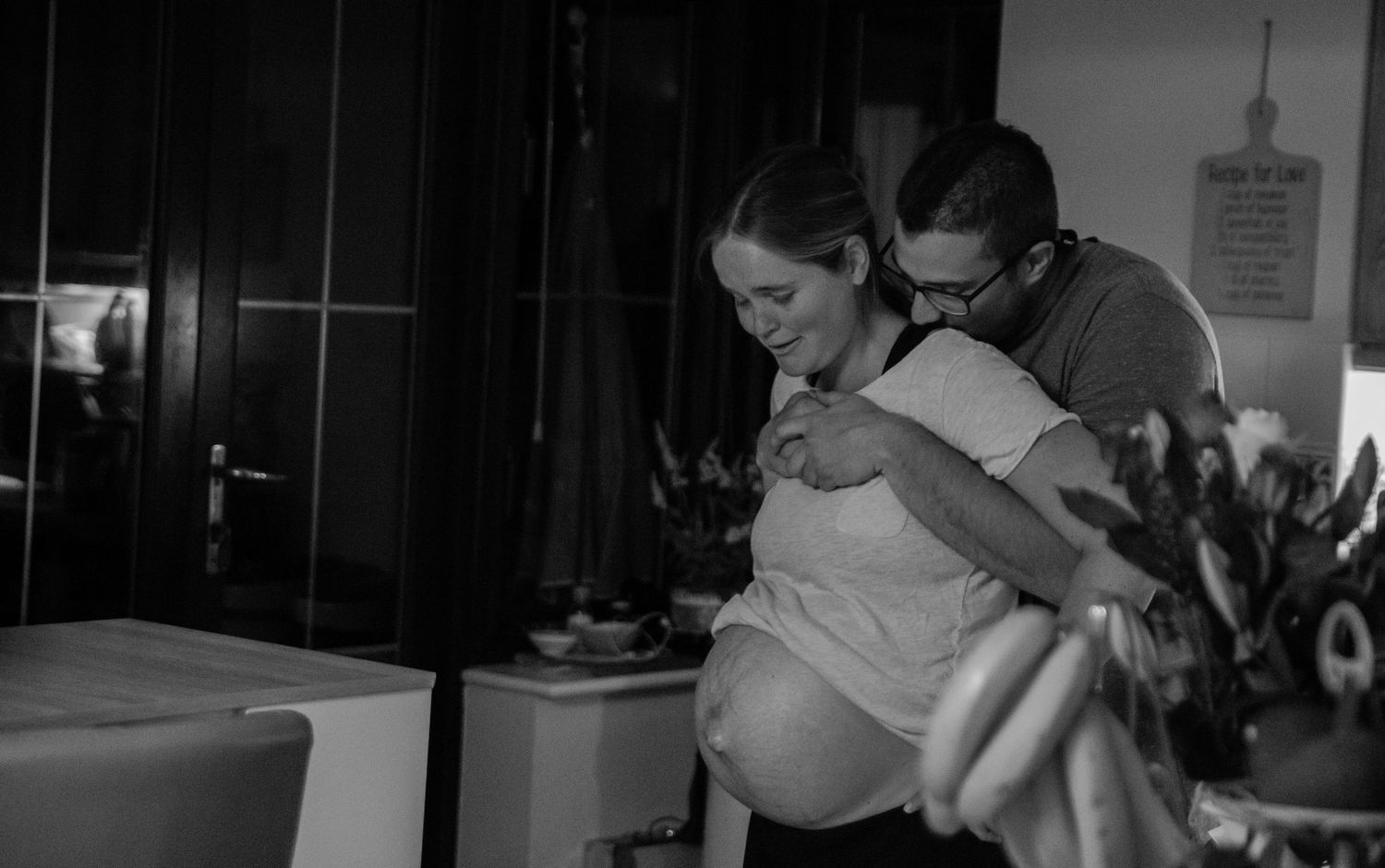 A black and white photo shows a pregnant woman leaning forward in labor while being supported from behind.