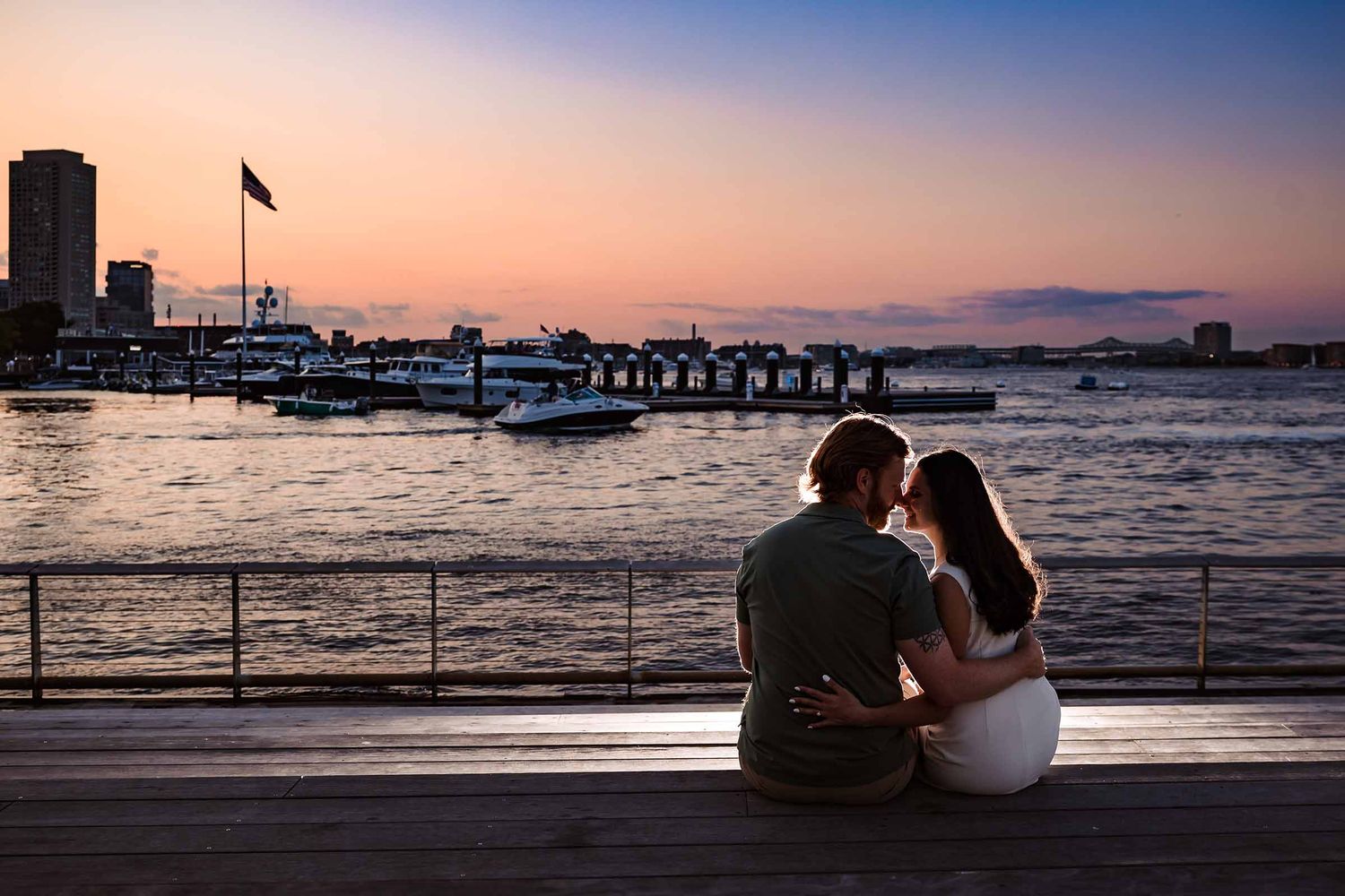 As the sunsets over Boston Harbor, the newly engaged couple share a kiss with the colorful sky as a backdrop,