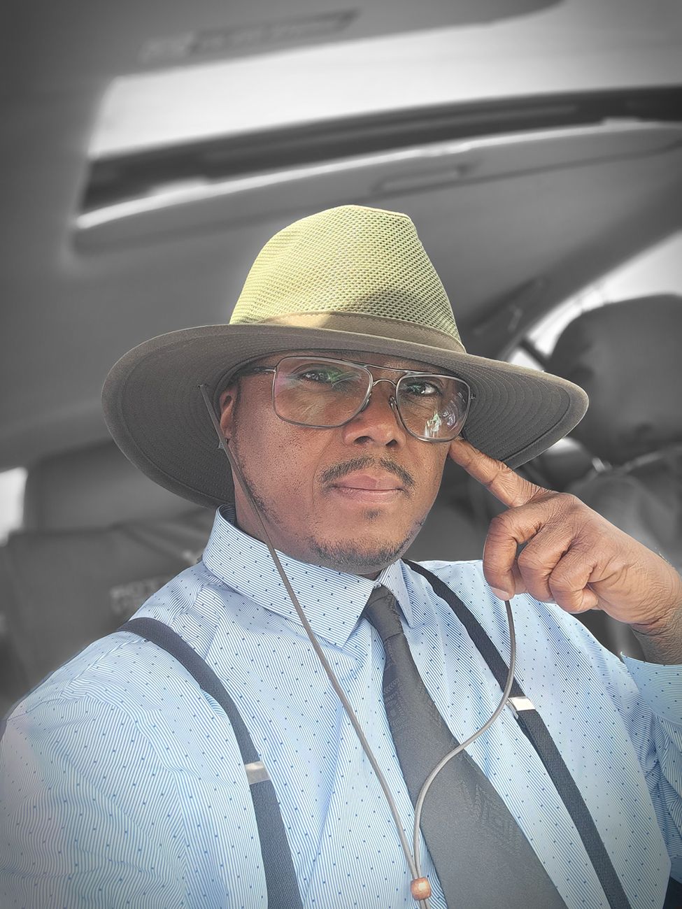 A person in business attire wearing a light colored fedora hat, suspenders and glasses sits in a vehicle.