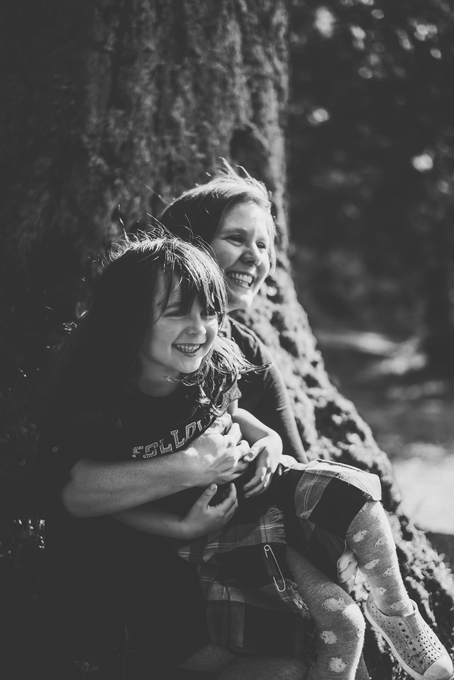Two people share a joyful moment while sitting against a large tree trunk.