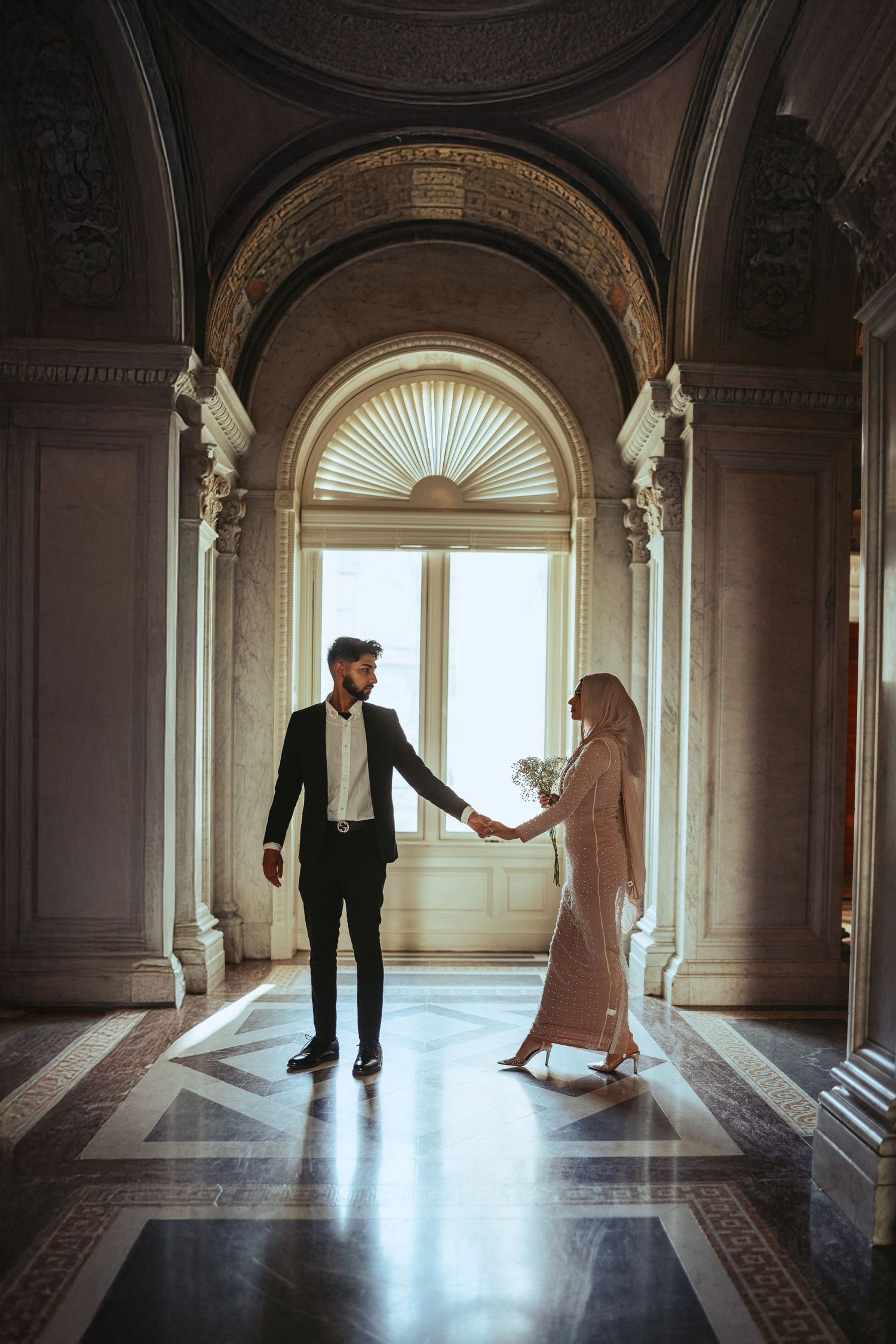 Two people hold hands in a grand hallway with arched ceilings, ornate columns and a sunburst window at the far end.