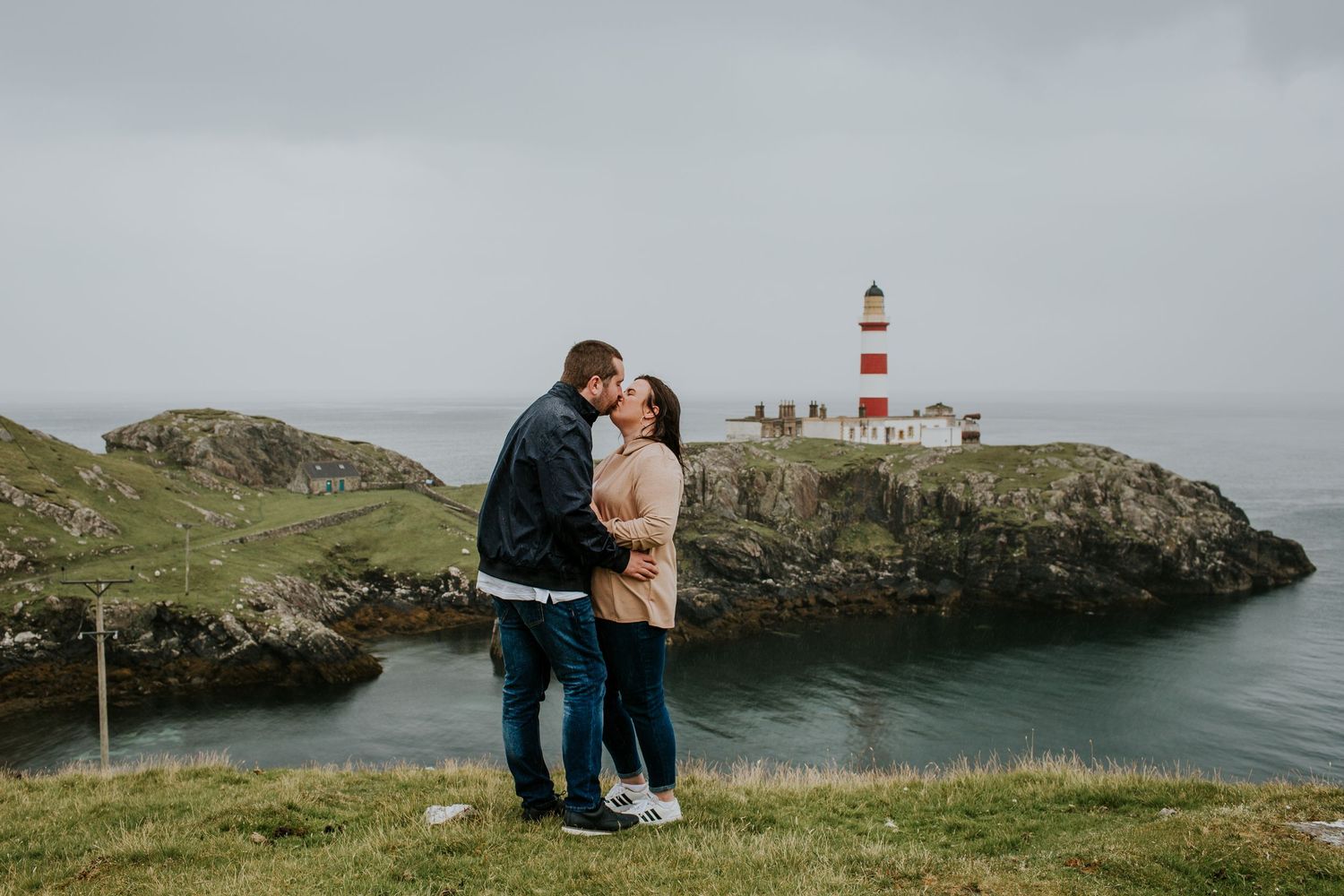 A couple embraces near a red and white lighthouse perched on dramatic coastal cliffs.
