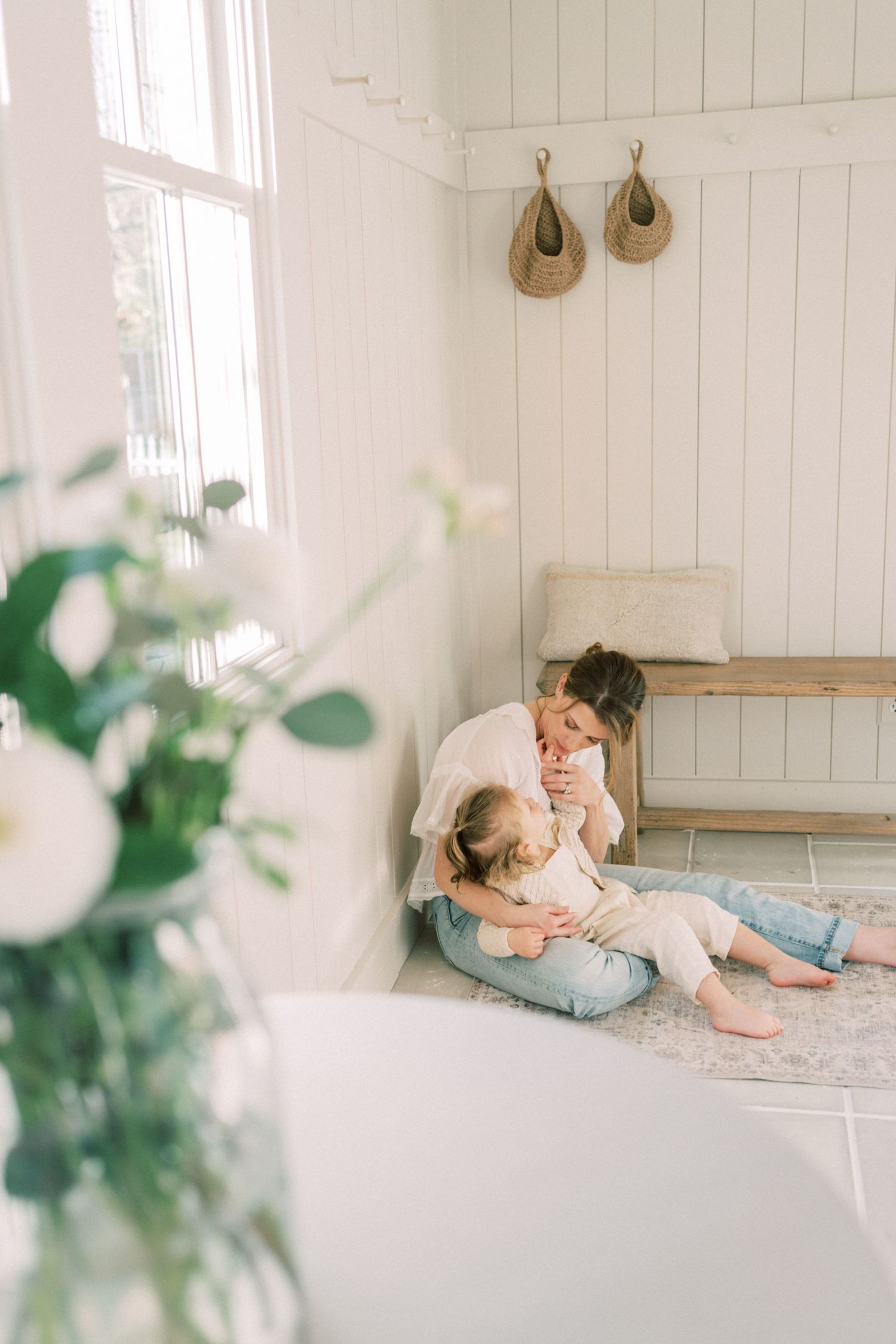 A bright, minimalist white room features hanging decor above a wooden bench and green plants in soft focus.