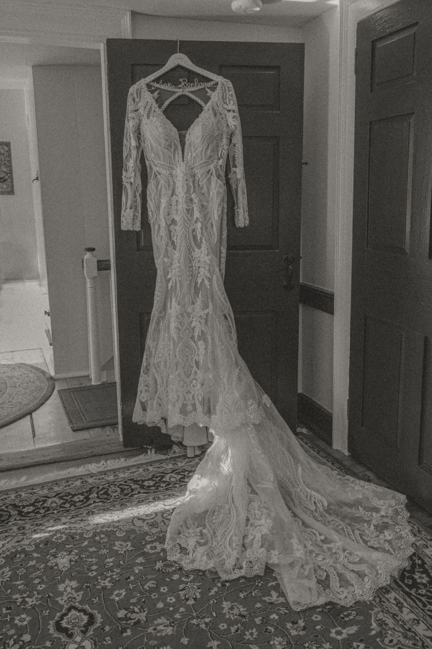 A vintage-style black and white photograph by Samantha Campbell of a lace wedding dress with long sleeves and train displayed on a hanger.
