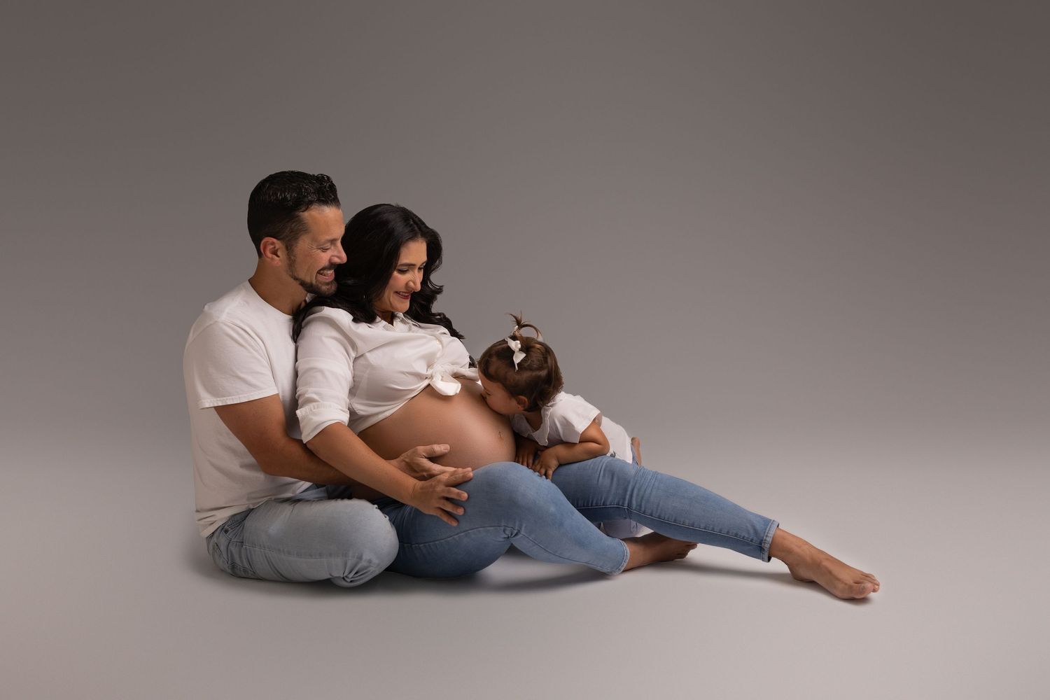A pregnant woman in flowing white fabric poses gracefully against a light gray background in an artistic tampa maternity photo.