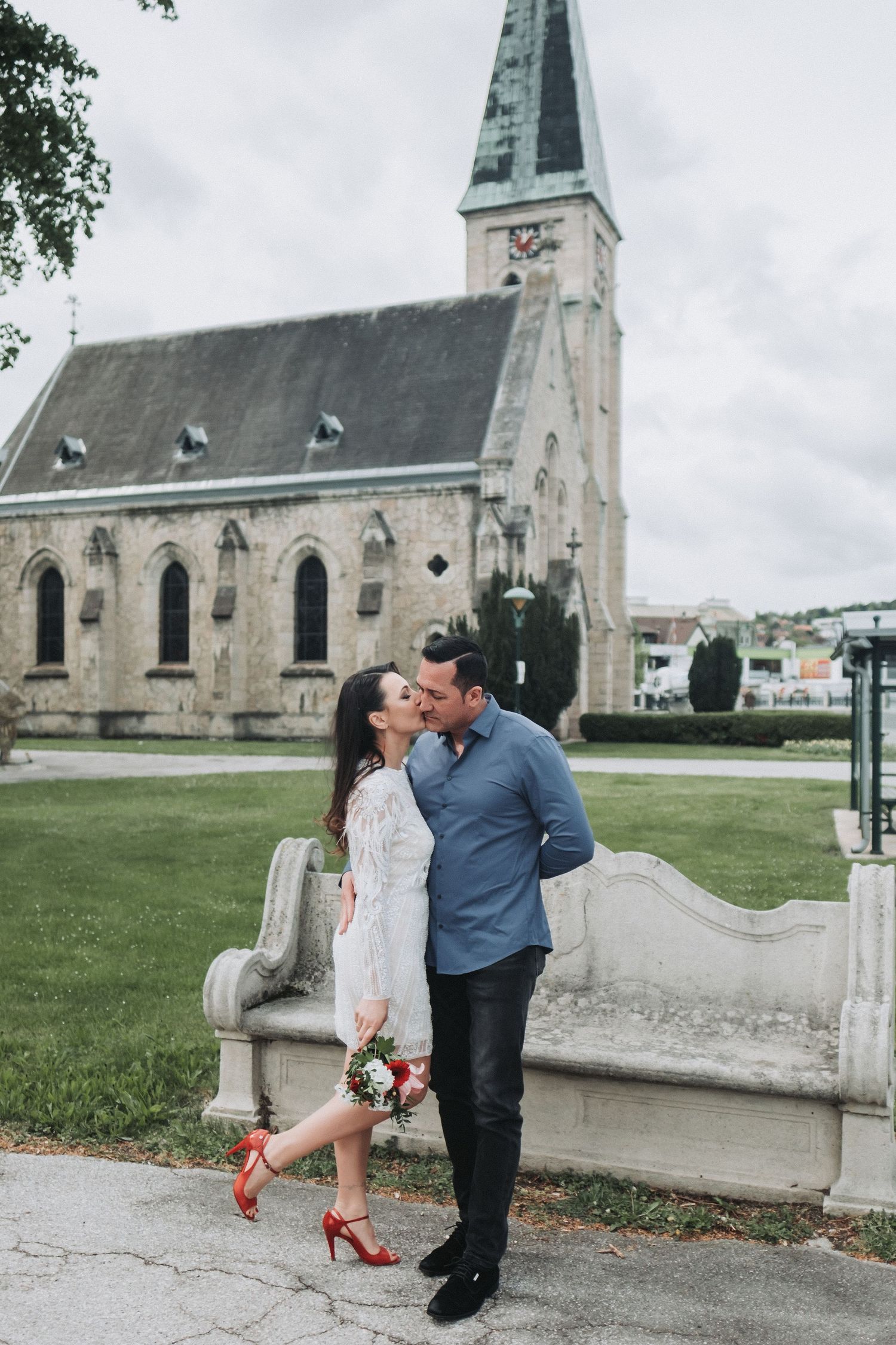 Romantic couple shares kiss outside historic church with red heels adding pop of color.