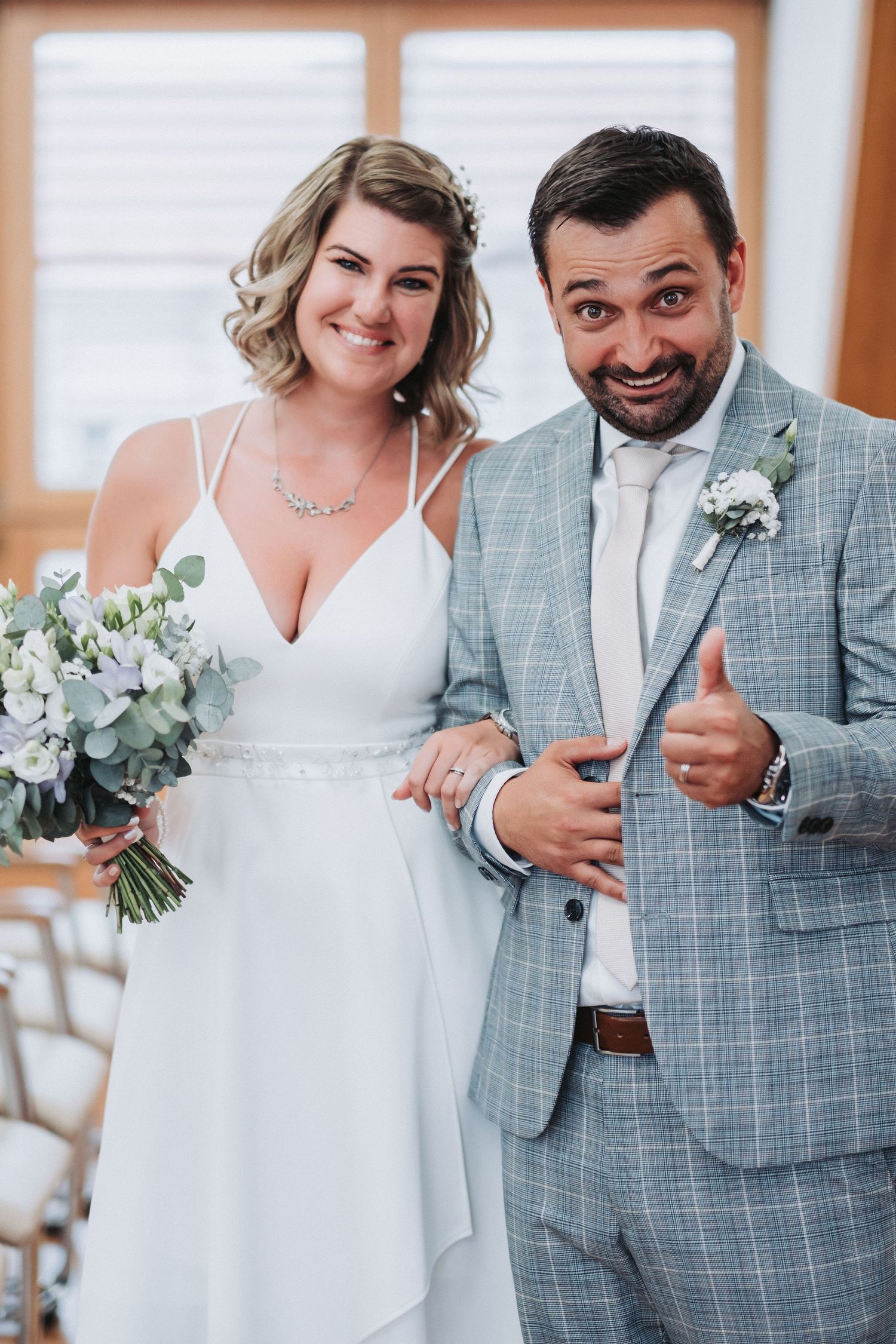 Happy newlyweds pose together, bride holding eucalyptus bouquet with groom in checkered suit.