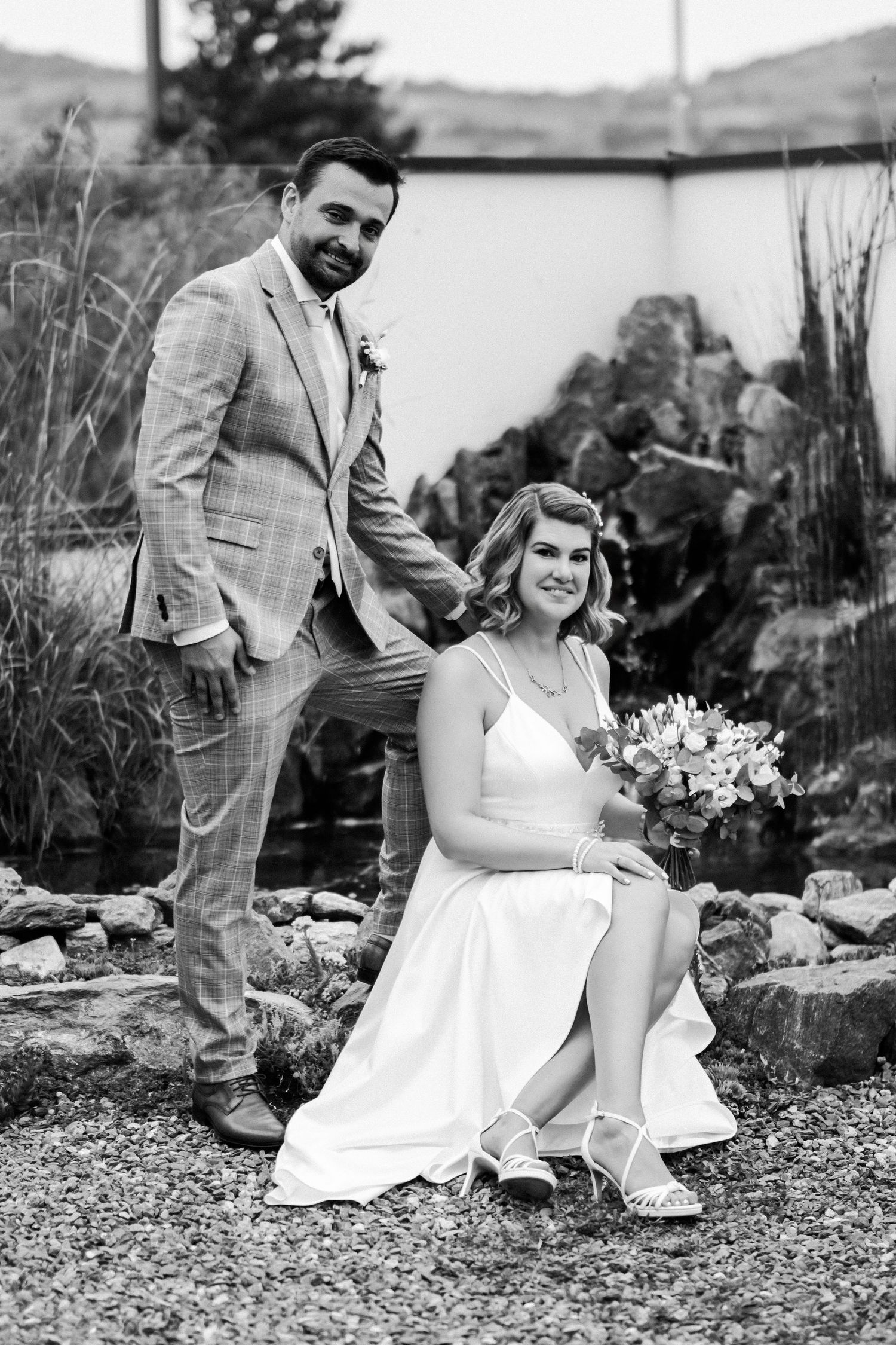 Outdoor black and white portrait of newlyweds in garden setting with bride seated on rocks.