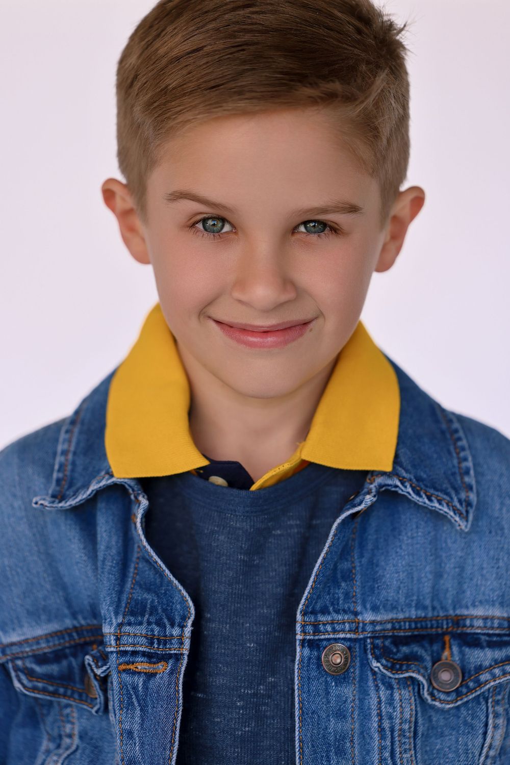 Studio portrait of a child in a denim jacket with a yellow collar smiling at the camera.