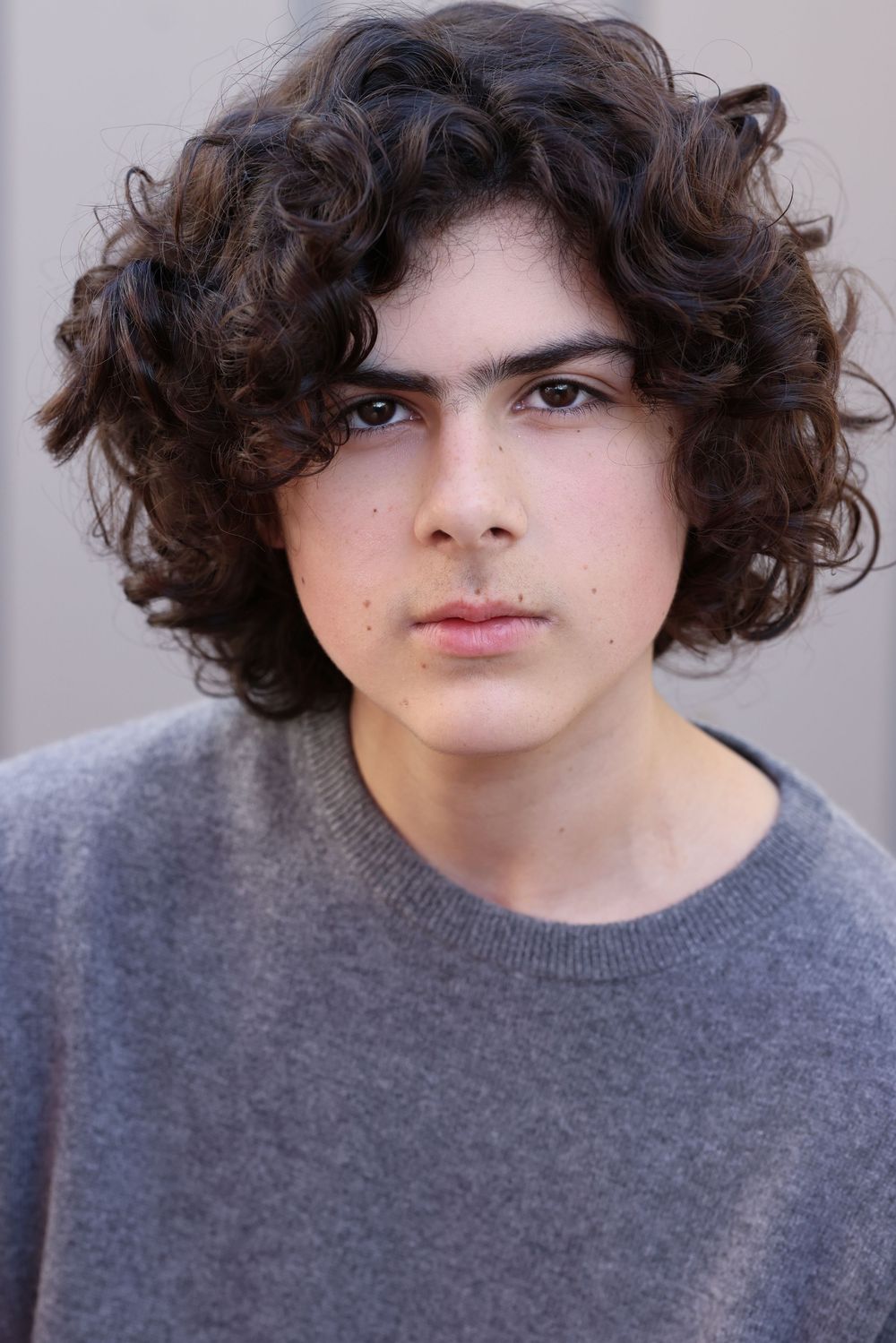 Portrait of curly-haired person in grey sweater against neutral background.