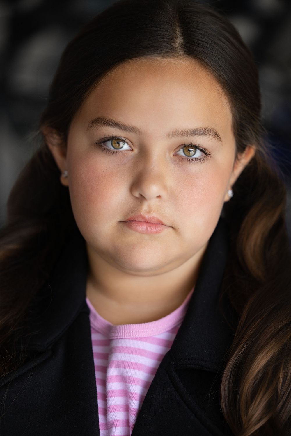 A portrait photo shows a person with long dark hair wearing a black jacket and pink striped shirt against a blurred background.