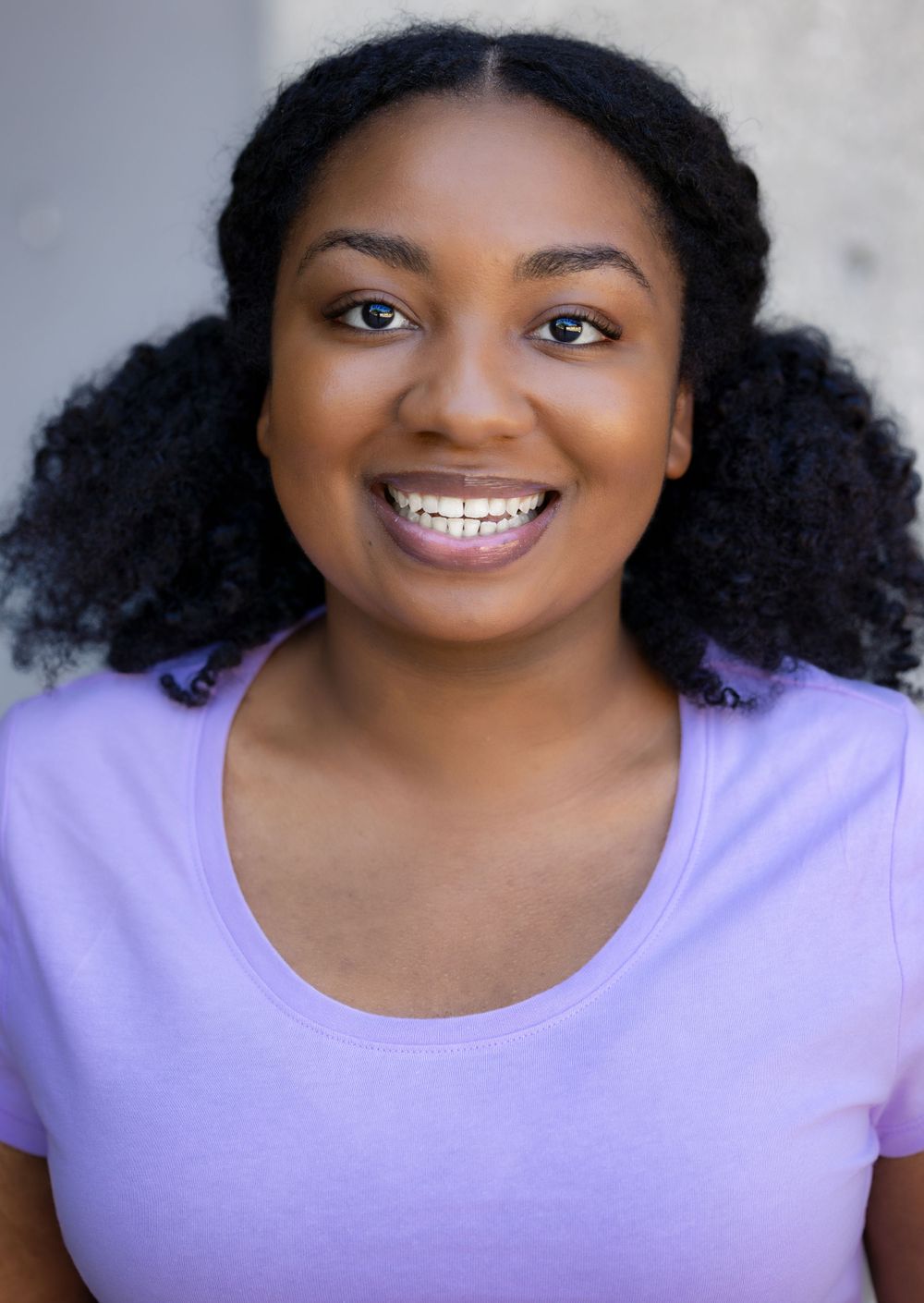 A person with curly hair in space buns wearing a purple t-shirt smiles brightly at the camera.