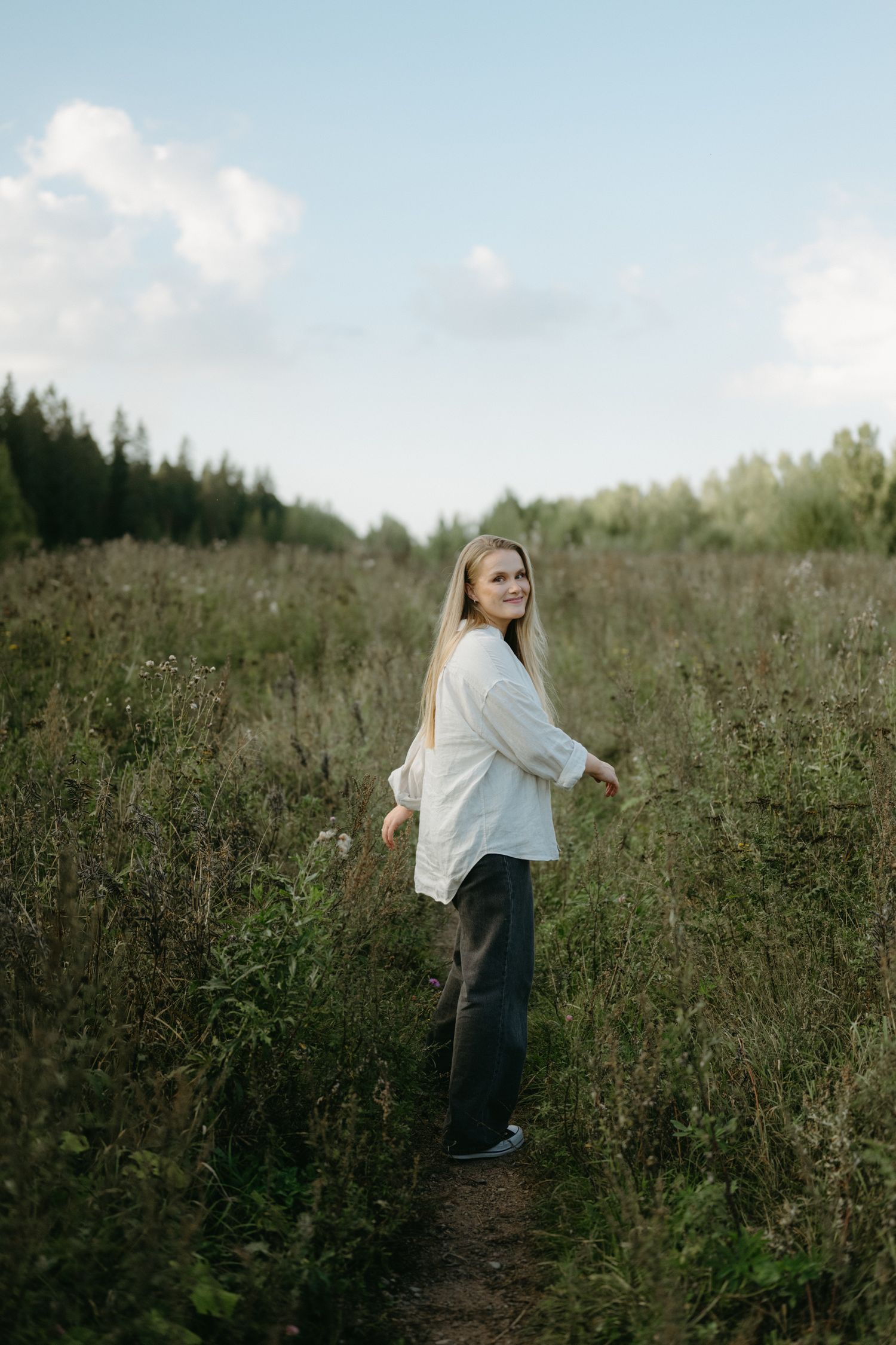 A person in a white sweater stands on a winding trail through a scenic meadow with evergreen trees.
