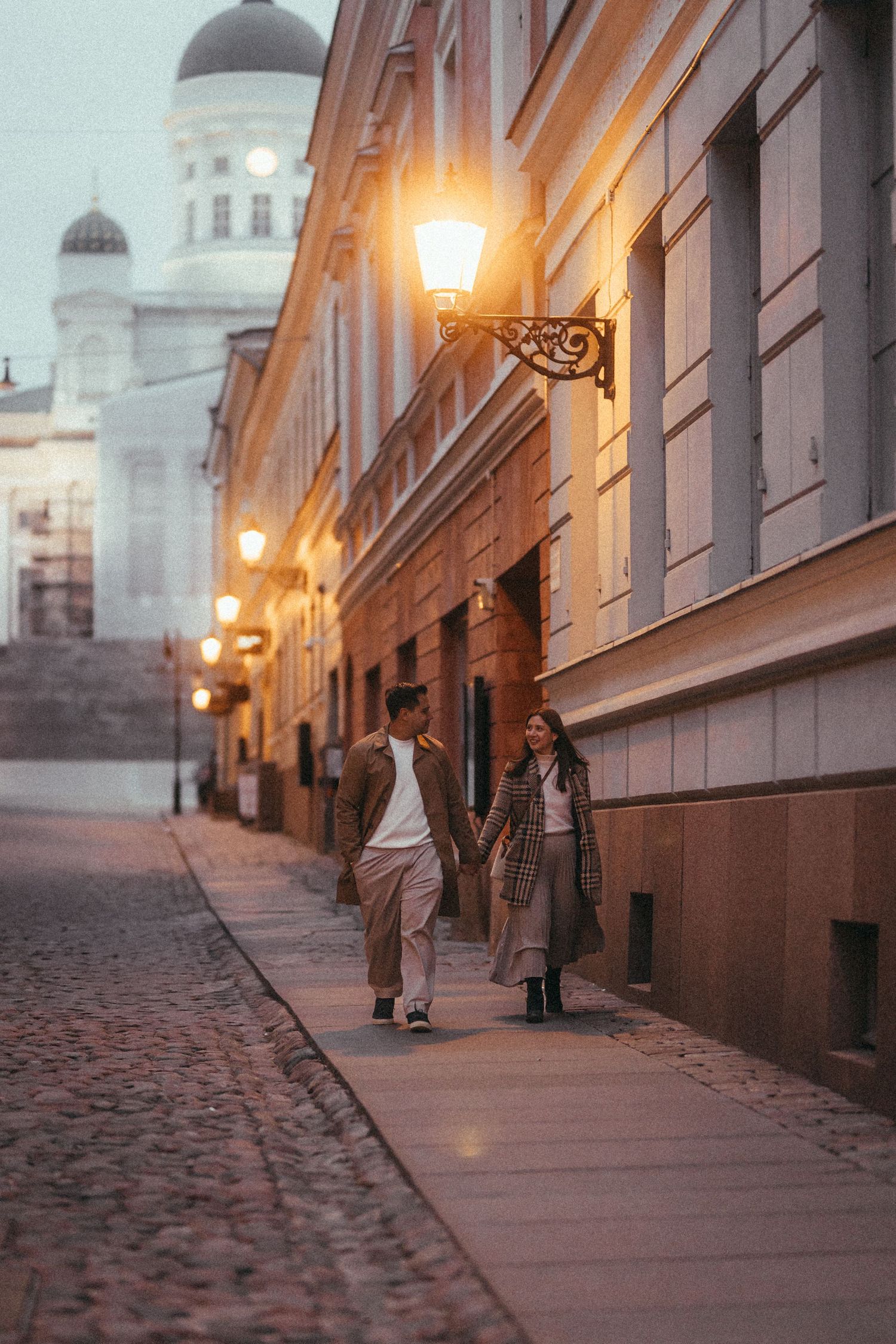 Couple walking down illuminated European street with cathedral dome in background at dusk.