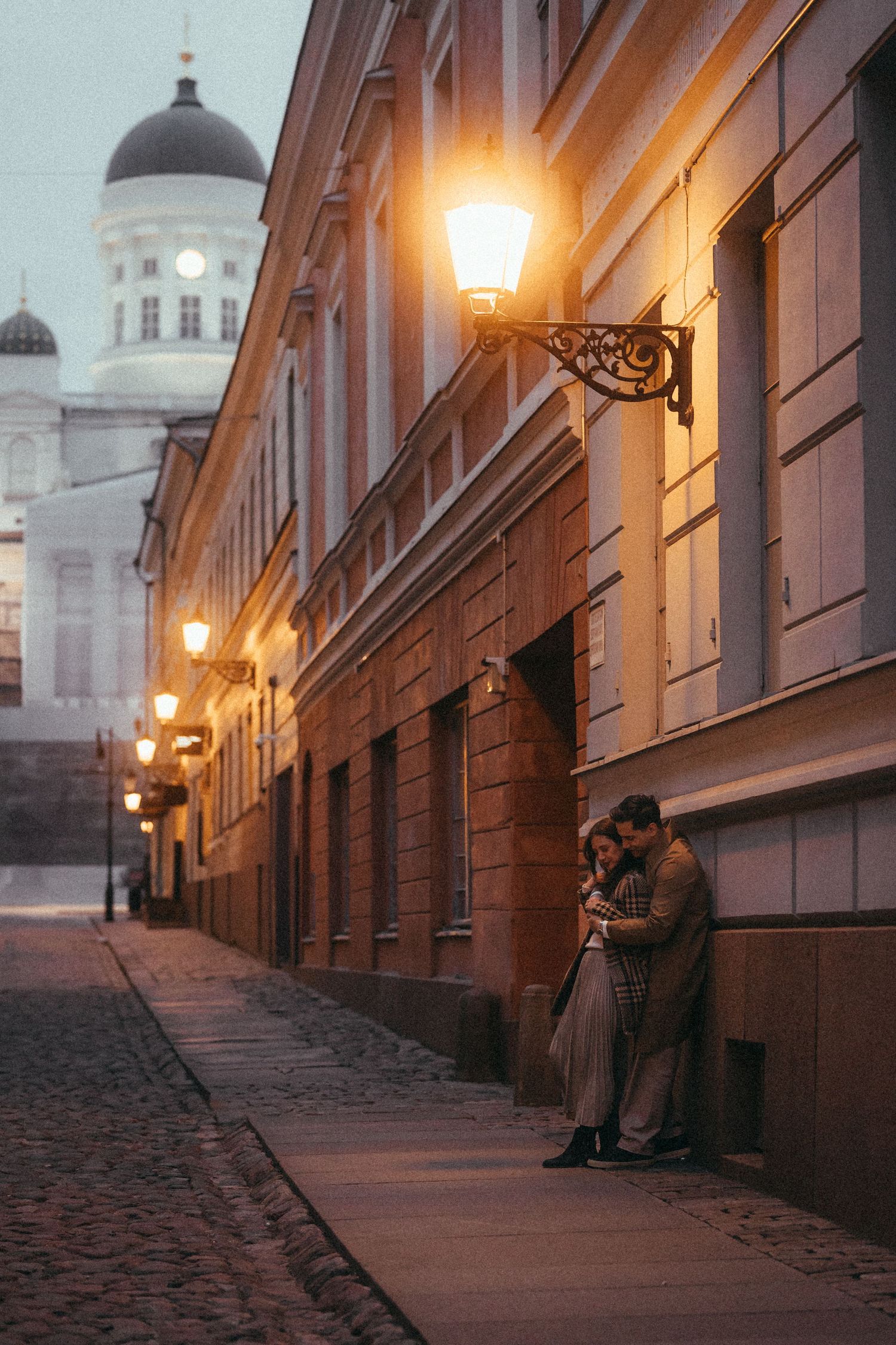 Couple embracing on cobblestone street with historic architecture and glowing street lamps.